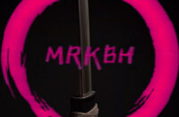 MRKBH stands in a dimly lit room as sacred symbols glow behind him, capturing the spiritual intensity of the Shattered Vessels single produced by Rico James.