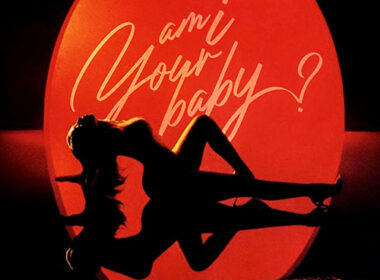 Young Lyric in a sultry scene from the "Am I Your Baby" music video, wearing lingerie and performing under moody lights.