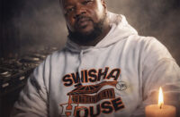 Photorealistic memorial tribute of DJ Michael “5000” Watts wearing a white Swishahouse hoodie, seated before DJ turntables with a candle glowing at his side, honoring the Houston hip-hop pioneer.