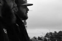lack-and-white image of two Hip-Hop artists, Legit and HostileProd, standing side by side on a city street, wearing caps and looking forward, capturing a gritty, cinematic underground Hip-Hop aesthetic