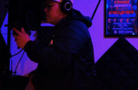 Hip-Hop artist recording in a dimly lit studio wearing headphones and holding a microphone, surrounded by acoustic panels and neon lighting, capturing a focused freestyle session atmosphere.
