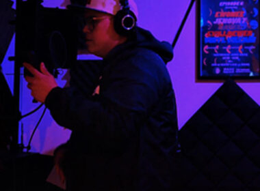 Hip-Hop artist recording in a dimly lit studio wearing headphones and holding a microphone, surrounded by acoustic panels and neon lighting, capturing a focused freestyle session atmosphere.