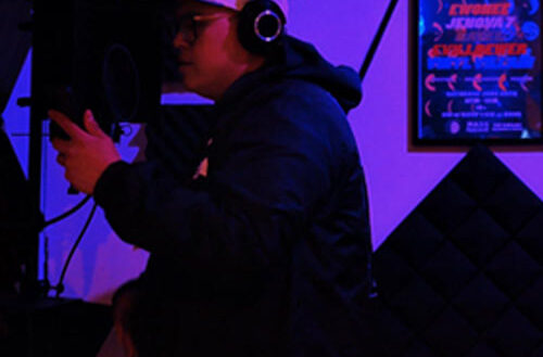 Hip-Hop artist recording in a dimly lit studio wearing headphones and holding a microphone, surrounded by acoustic panels and neon lighting, capturing a focused freestyle session atmosphere.