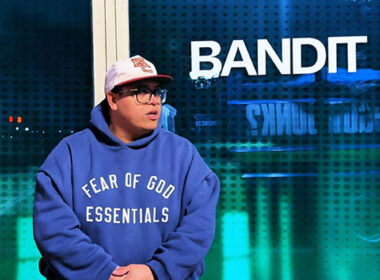 Hip-Hop artist wearing a hoodie stands near a window at night with city lights in the background, creating a reflective mood for the “So We Loved Ourselves” visual.