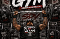 Street Da Villan bench presses oversized speaker weights on the cover of his single "In The Gym," produced by Narrator Beats Incorporated, symbolizing the discipline required to master bars, flow, delivery, and lyricism.